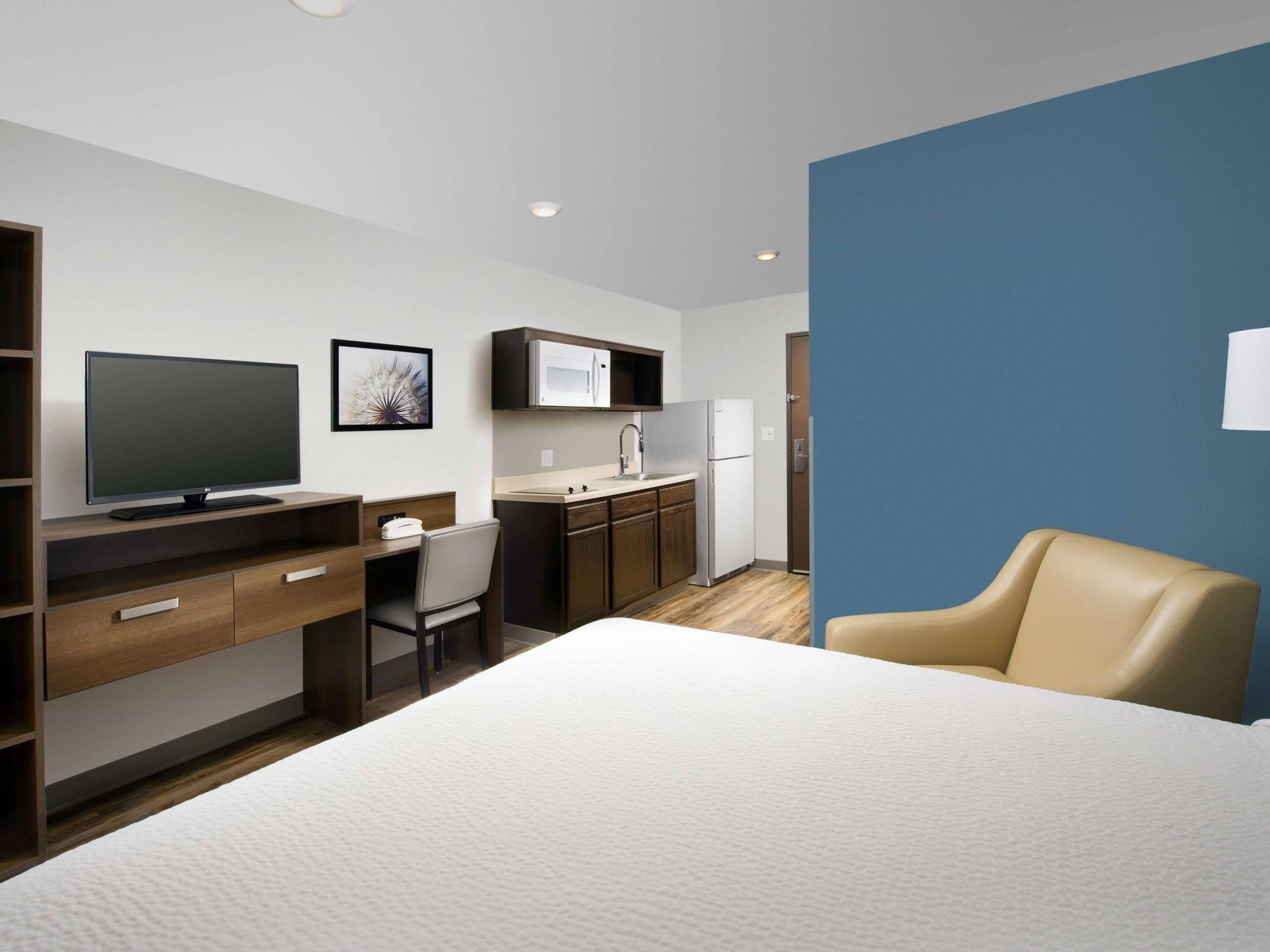 Habitación de hotel con una cama, un escritorio con computadora, una silla, un televisor y una kitchenette con microondas y lavamanos.