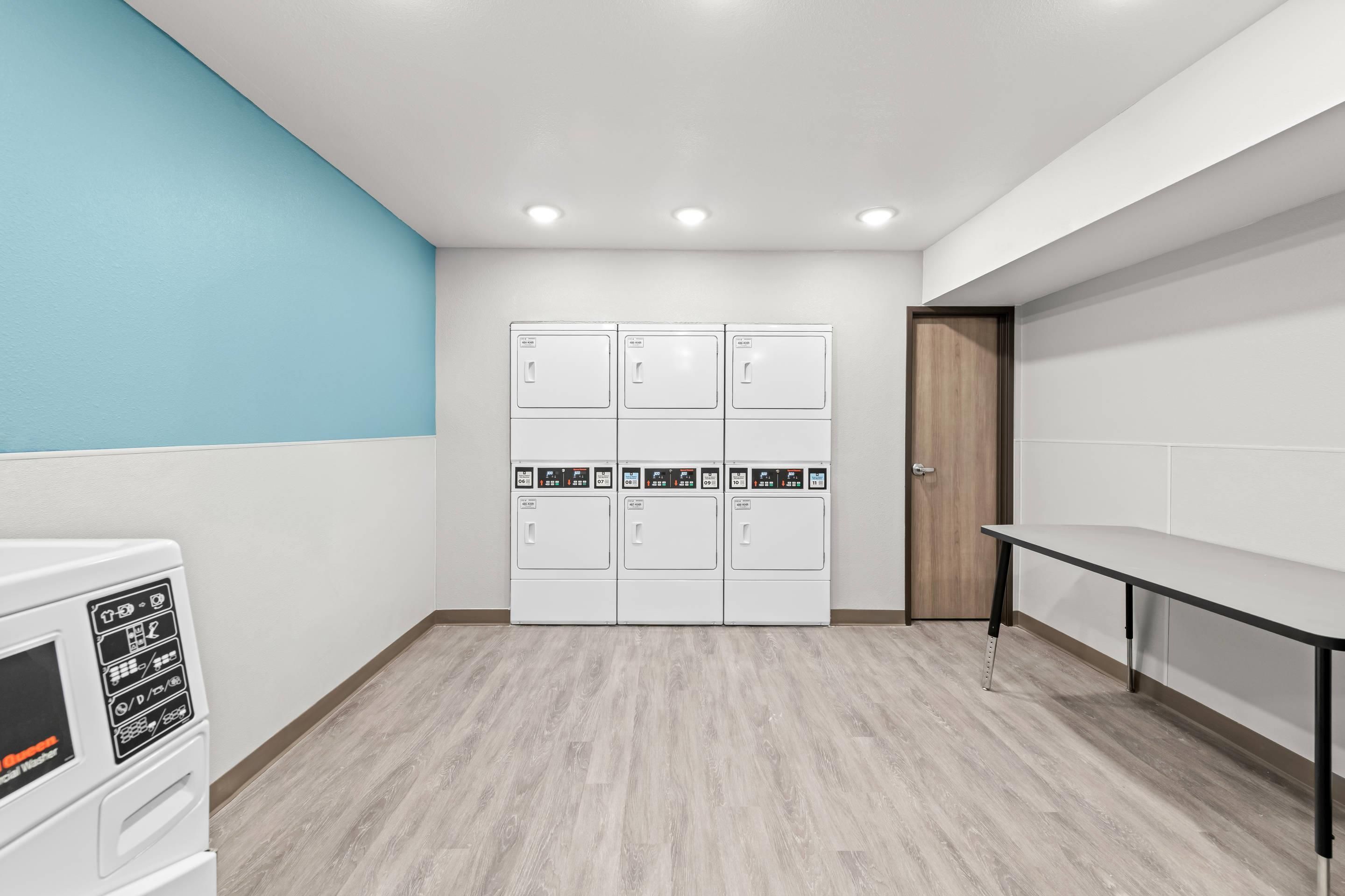 Un cuarto de lavandería con una hilera de lavadoras apiladas, una puerta de madera y un banco.