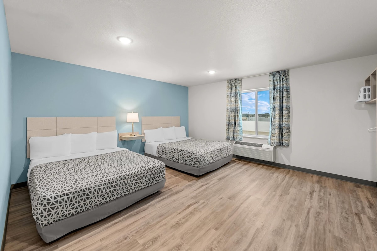 A hotel room with two beds, a nightstand, a lamp, a window with curtains, and an air conditioning unit.