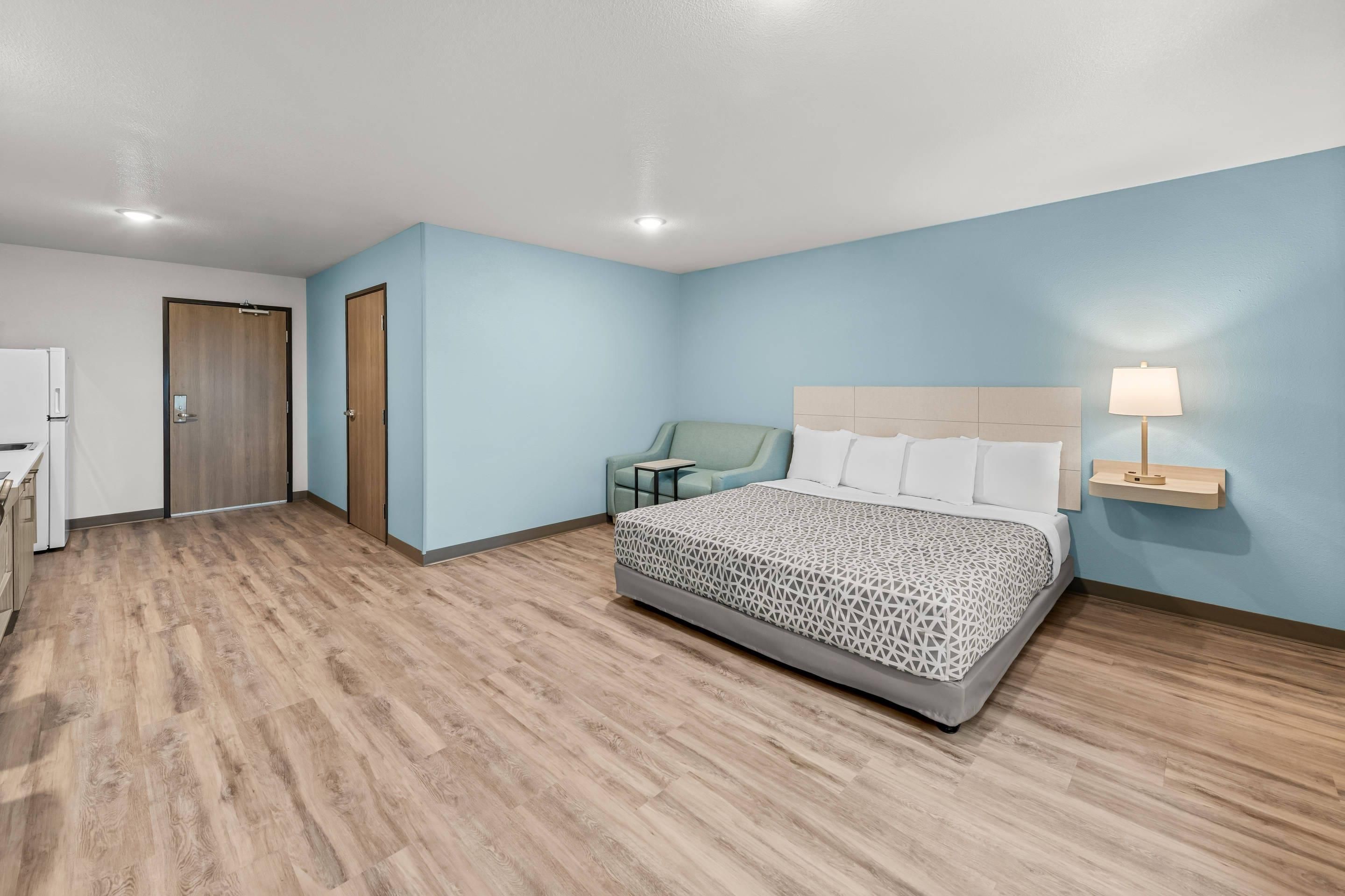 A hotel room with a bed, a desk, a chair, and a nightstand with a lamp.