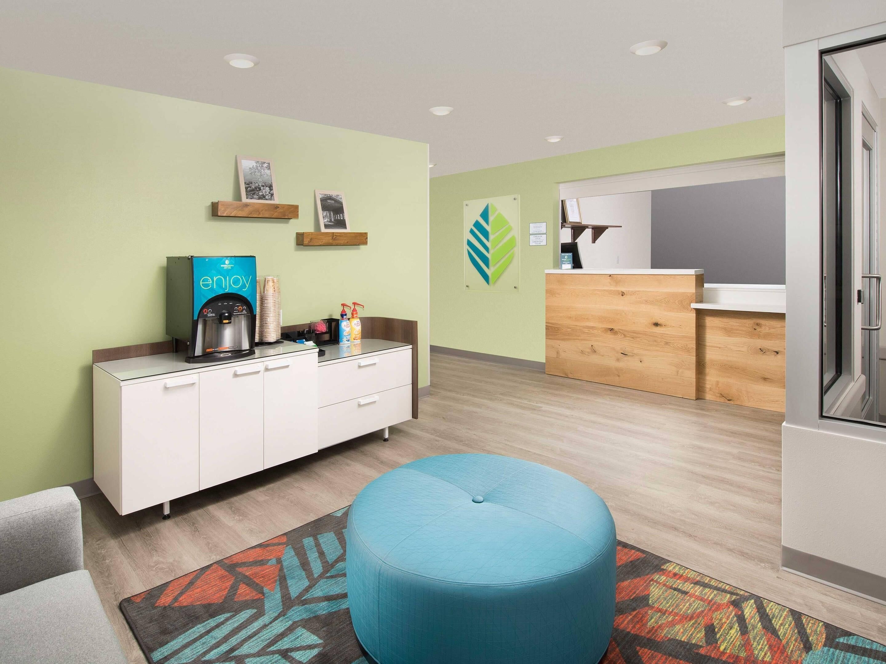 A modern hotel room with a kitchenette, a blue ottoman, and a patterned rug.