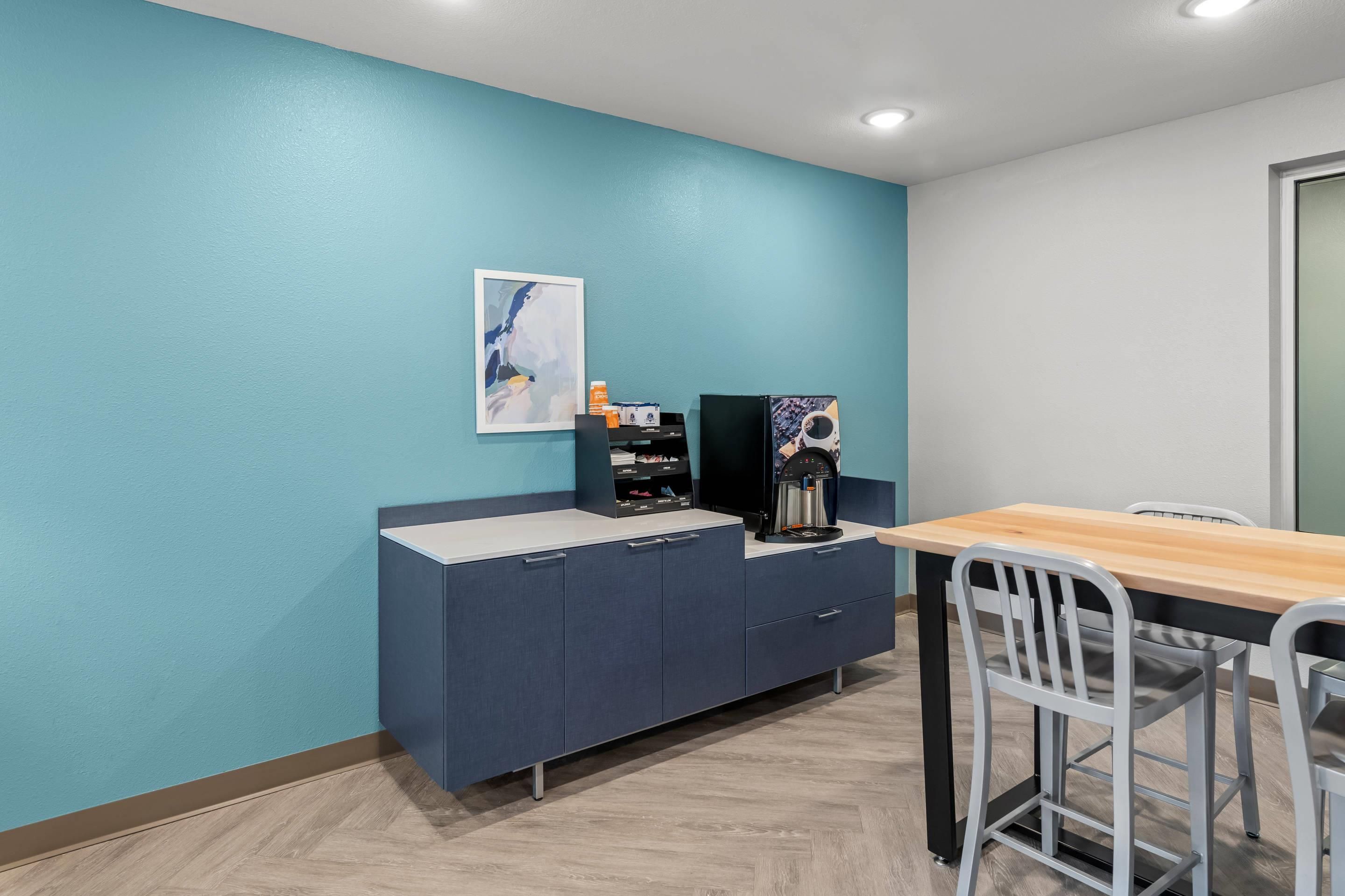 Une pièce avec un mur de couleur turquoise, une table en bois, une chaise blanche, un meuble bleu foncé et une machine à café.
