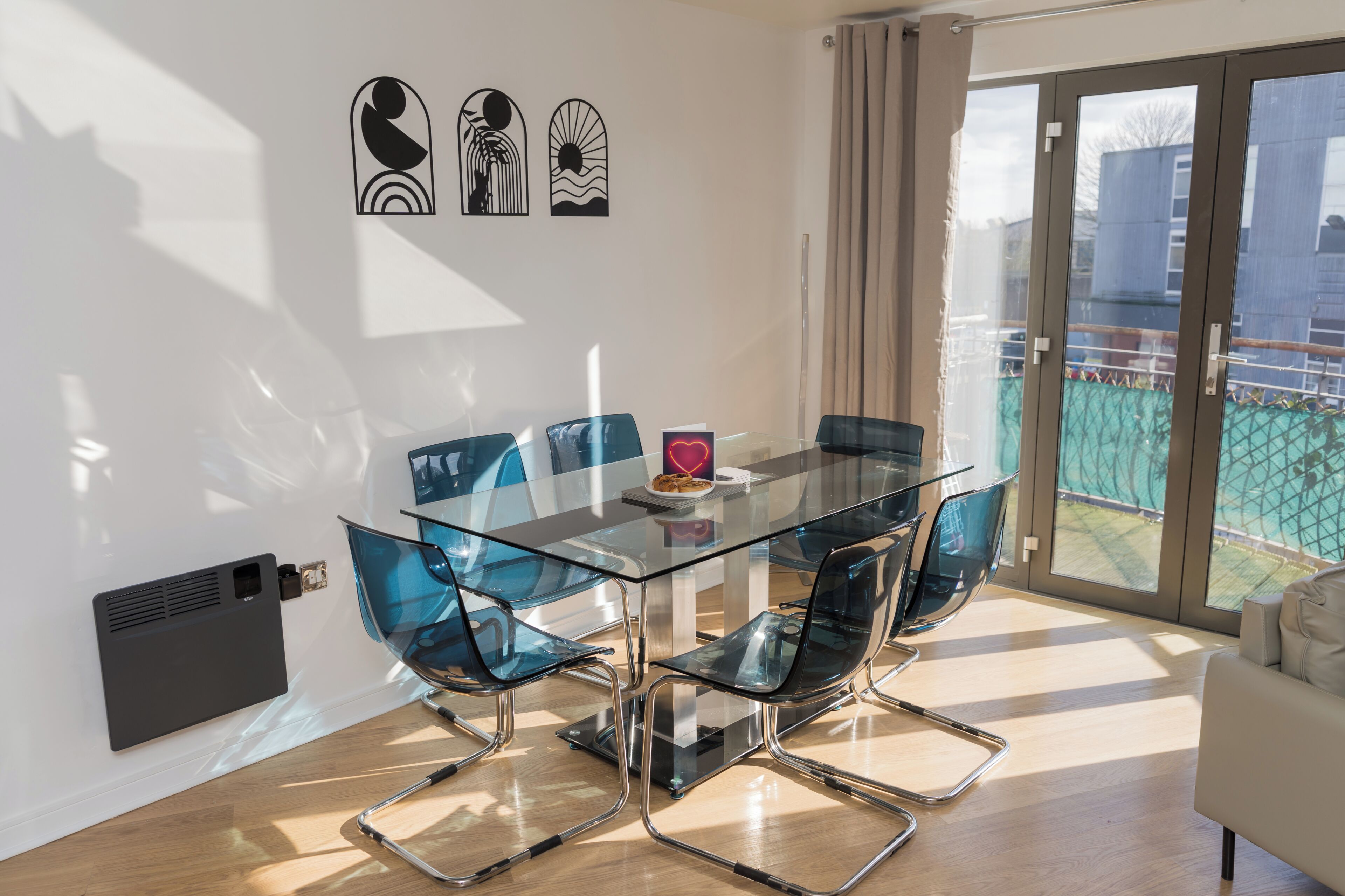 A modern dining area with a glass table and metal chairs, a white wall with artwork, and a sliding glass door to a balcony.