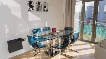 A modern dining area with a glass table and metal chairs, a white wall with artwork, and a sliding glass door to a balcony.