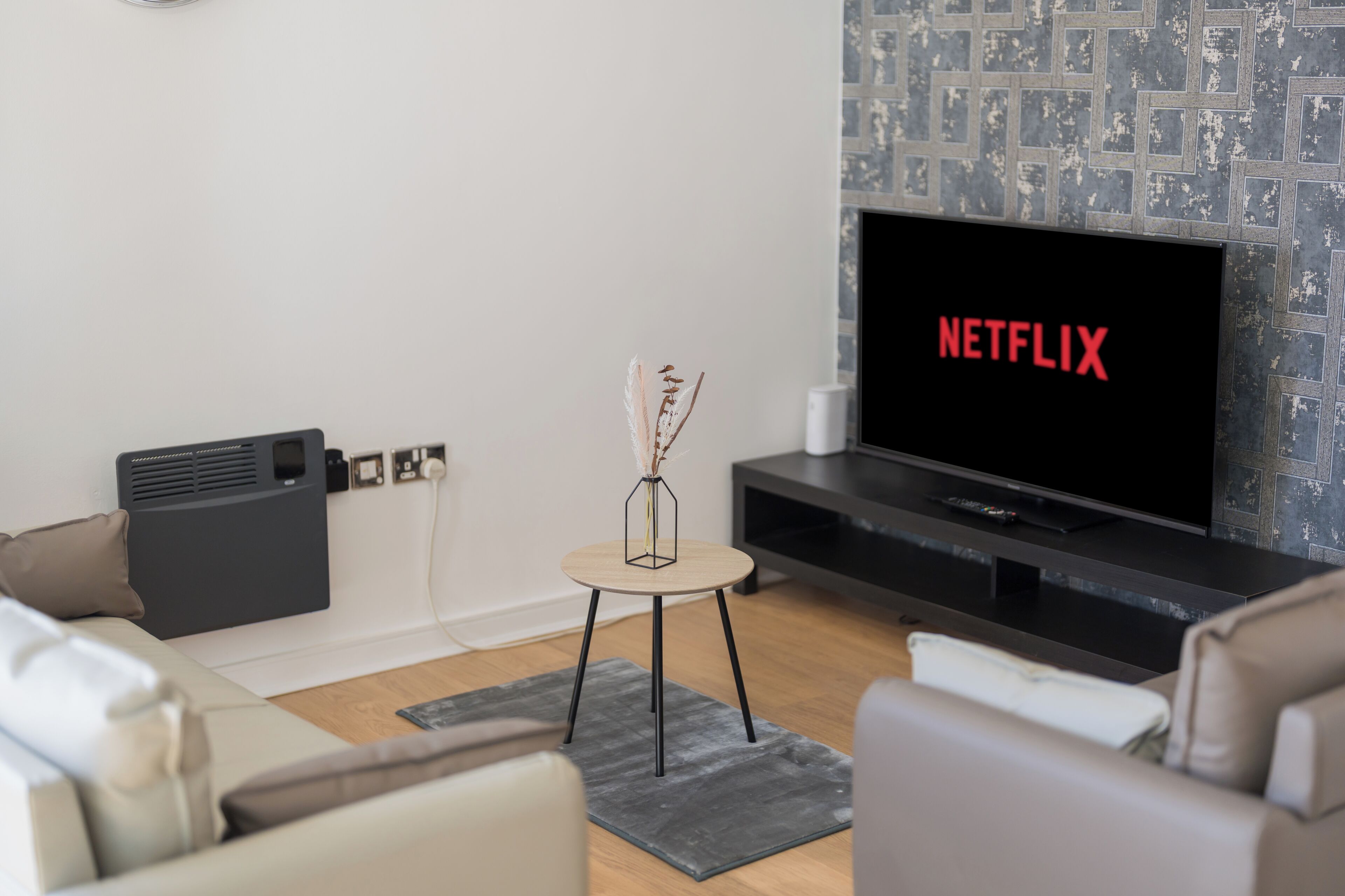 A modern living room with a flat-screen TV displaying 'NETFLIX' in red letters, a small side table with a vase, and a grey patterned wall.
