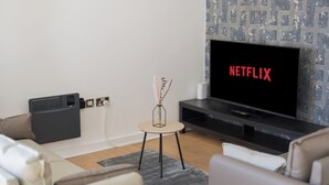 Moderni olohuone, jossa on litteänäyttöinen televisio, jonka ruudulla näkyy punaisilla kirjaimilla teksti »NETFLIX», pieni sivupöytä, jossa on maljakko, ja harmaa, kuvioitu seinä.