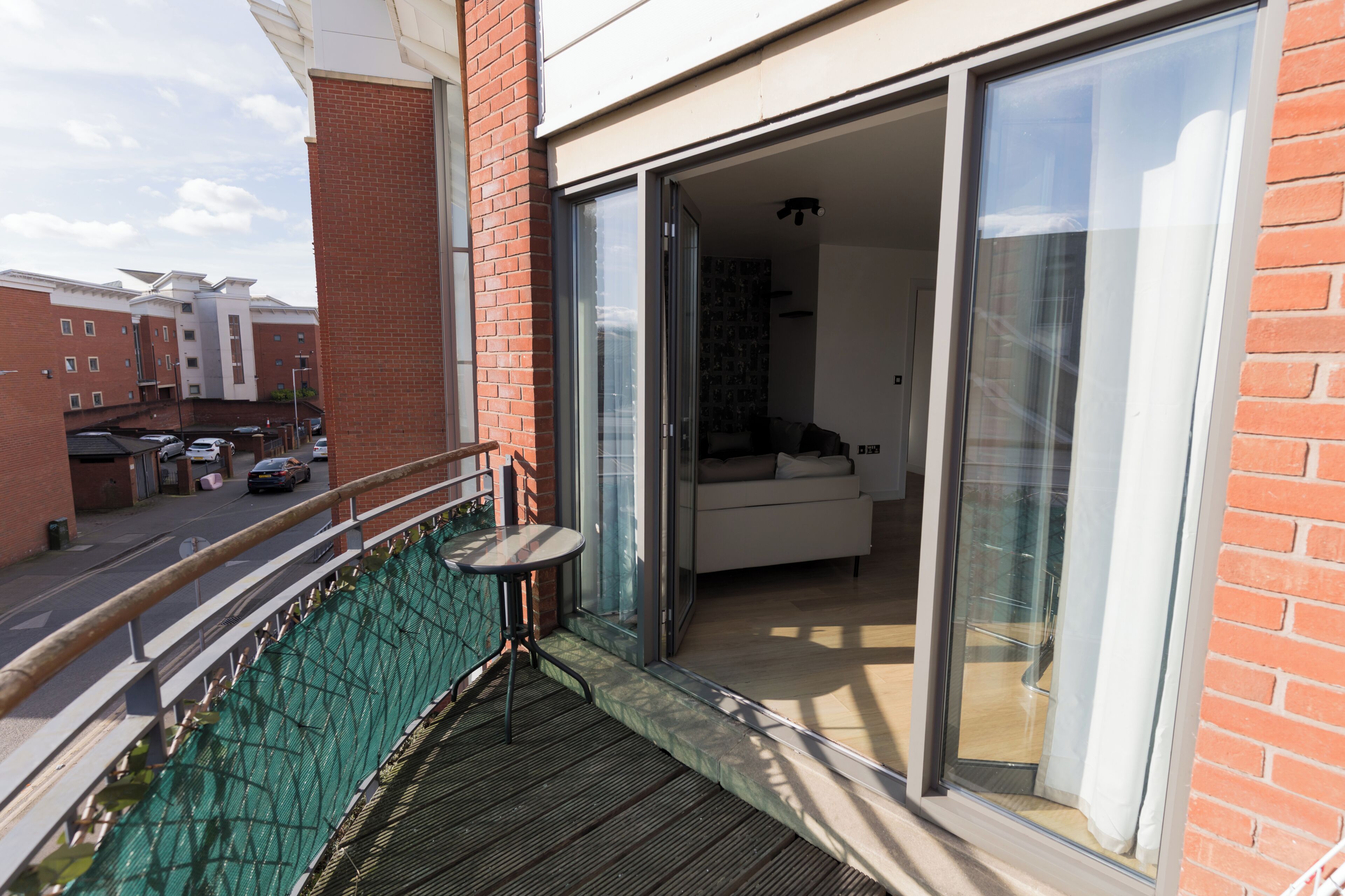 A balcony with a table and chairs, a sliding glass door leading to a living area, and a view of the street and other buildings.