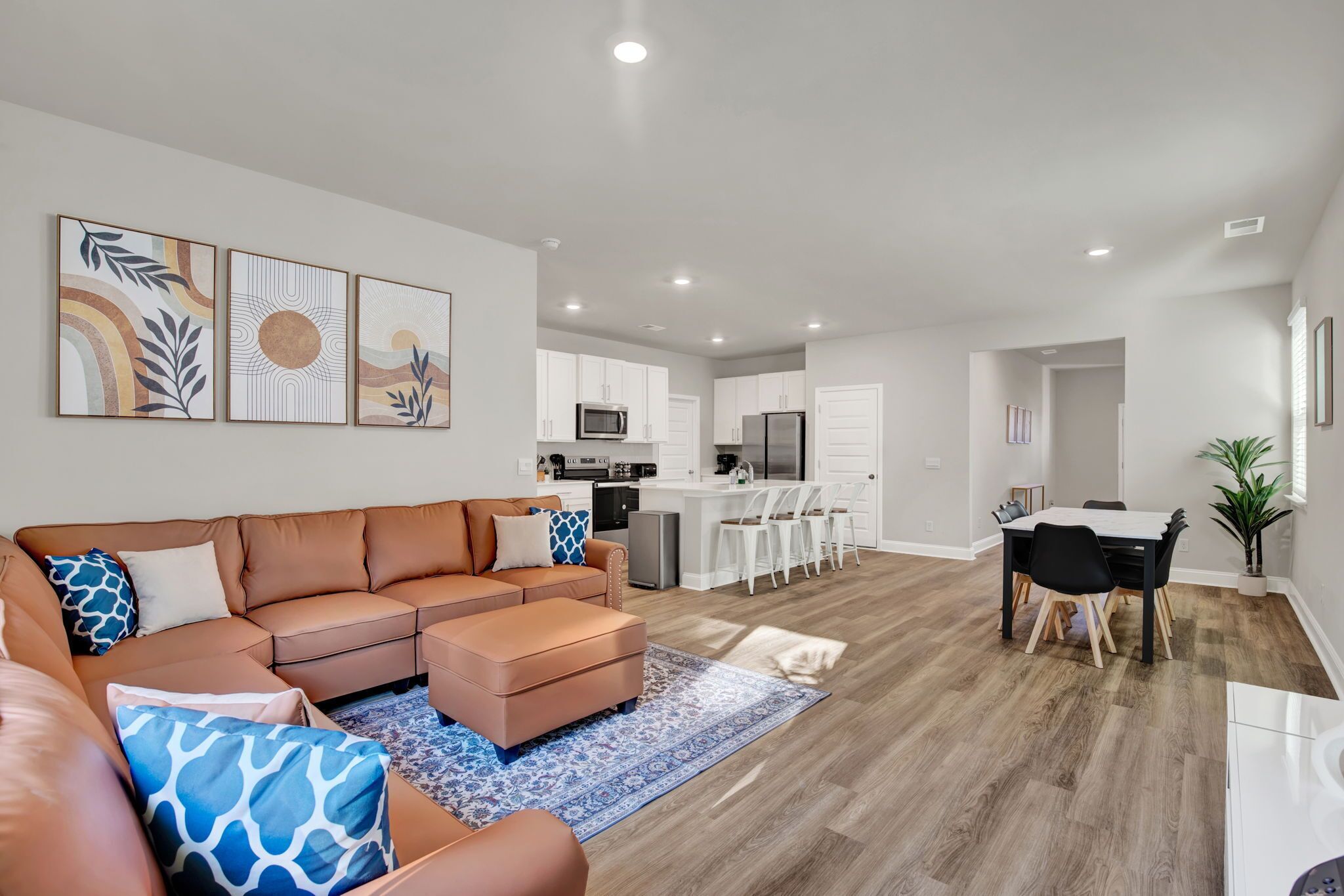 A modern living room with a brown sectional sofa, a dining area with a white table and chairs, and a kitchen with white cabinets and appliances.