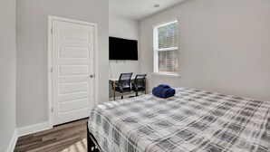 Quarto com cama, escrivaninha com duas cadeiras, TV fixada na parede e janela com persianas.