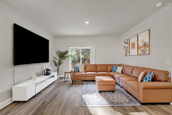 Un salon moderne doté d’un grand canapé d’angle, d’un téléviseur à écran plat, d’un meuble de rangement blanc et d’une grande fenêtre donnant sur la verdure.