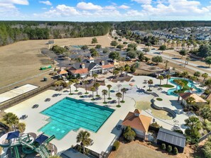 Vue aérienne d’un quartier résidentiel avec une grande piscine, plusieurs maisons et un parc équipé d’aires de jeux pour enfants.
