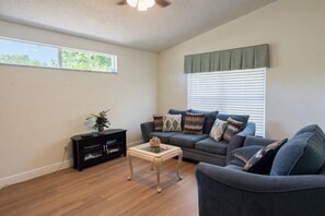 A living room with a sofa, a coffee table, a TV stand, and a window with blinds. - 3BR Pool Home in Kissimmee Sleeps 6 (Kissimmee)