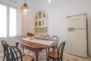 A dining area with a wooden table, chairs, a bowl of salad, a white refrigerator, and a window with blinds. - TD Palazzo Fiume 1 – Heart of Polignano (Polignano a Mare)