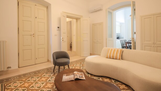 Ein Wohnzimmer mit einer Couch, einem Sessel, einem Couchtisch und einem gemusterten Teppich. Weiße Türen und eine Klimaanlage sind an der Wand angebracht.