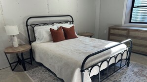 Uma cama bem arrumada com estrutura de metal, uma mesa de cabeceira com abajur e um penteado em um quarto com tapete estampado.