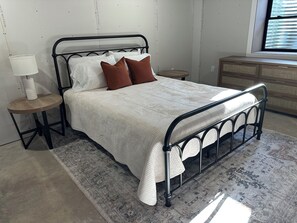 Una cama bien tendida con estructura metálica, una mesita de noche con lámpara y un aparador en una habitación con alfombra estampada.