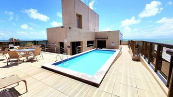 Uma área de piscina no terraço com vista clara para o mar, um espaço de descanso com cadeiras e um edifício com uma janela pequena.