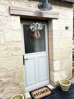 A stone house with a white door, a hanging flower wreath, a 'HOME' mat, and a potted plant on the porch. - Sunshine cottage2 (Fontevraud)