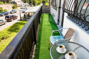 Een balkon met een groen kunstgrasveld, twee rieten stoelen en een tafel met twee koffiekopjes.