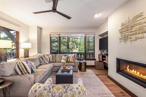 Ein Wohnzimmer mit Ecksofa, Kamin, Deckenventilator und großen Fenstern.