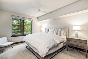 Ein Schlafzimmer mit einem Bett, Nachttischen, einem Sessel, einem Fenster mit Blick auf Bäume und einem Deckenventilator.