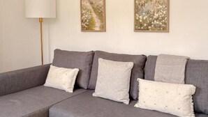 Ein graues Ecksofa mit drei Kissen vor einer schlichten Wand, eine Stehlampe links und zwei gerahmte Landschaftsbilder darüber.