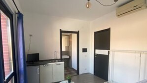 Quarto moderno com cama, uma pequena mesa branca e vista para uma cozinha e banheiro através de uma porta aberta.