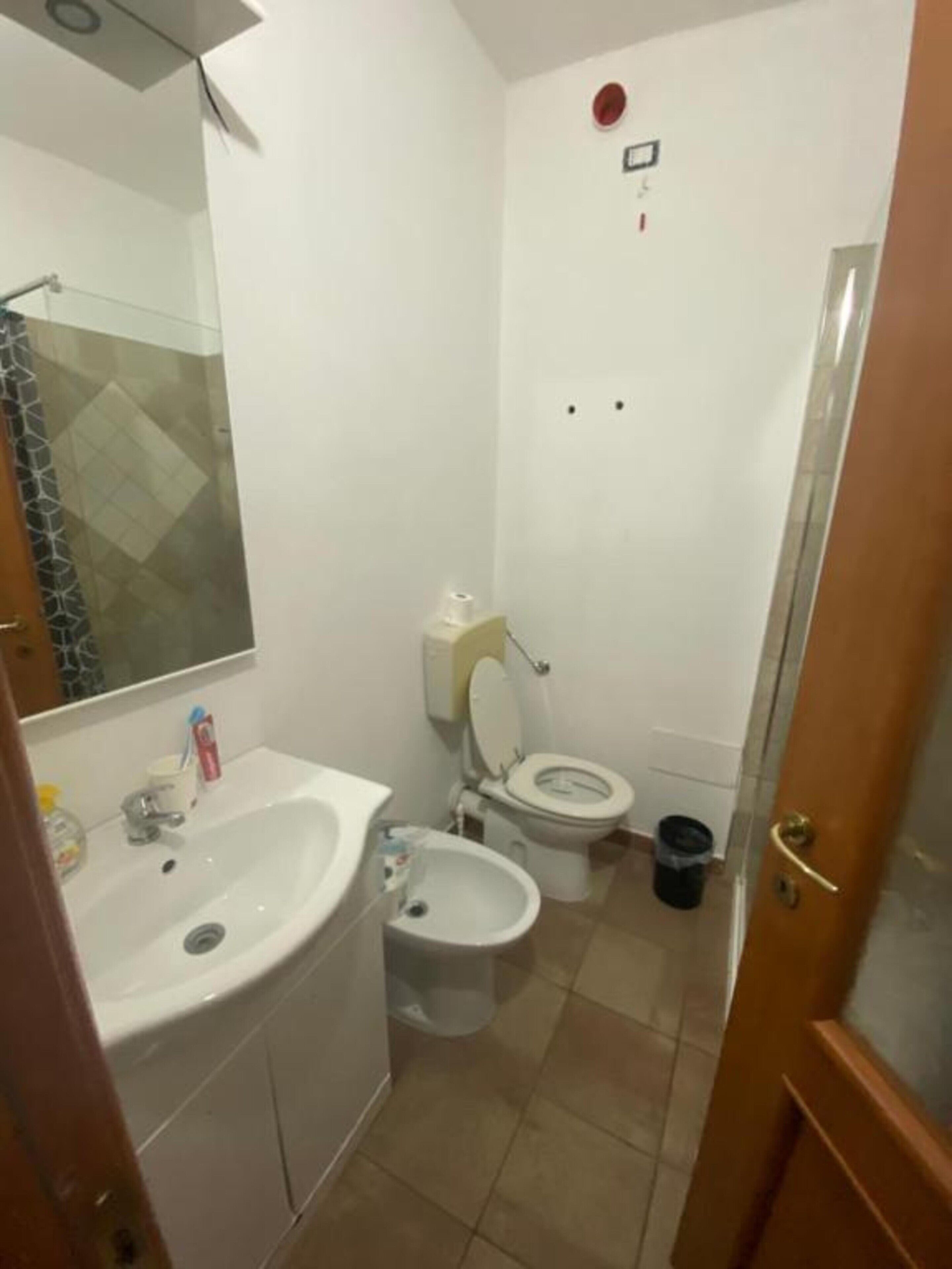 Een kleine badkamer met een wastafel, toilet en bidet. Er hangt een spiegel boven de wastafel en aan de rechterkant is een douchehoek.