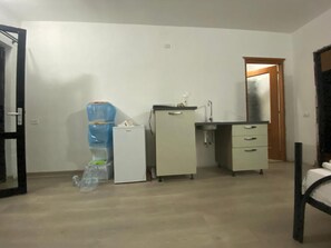 Ein Zimmer mit einem Bett, einer kleinen Küchenzeile, einem Wasserspender und einer Tür.