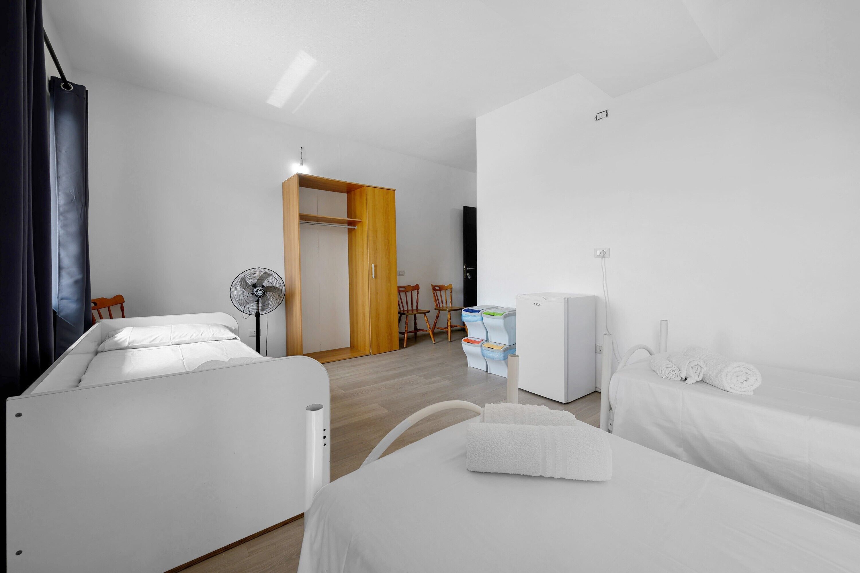 Een hotelkamer met twee bedden, een ventilator, een kledingkast, een klein tafeltje met een stoel en een witte radiator.