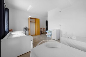 Een hotelkamer met twee bedden, een ventilator, een kledingkast, een klein tafeltje met een stoel en een witte radiator.