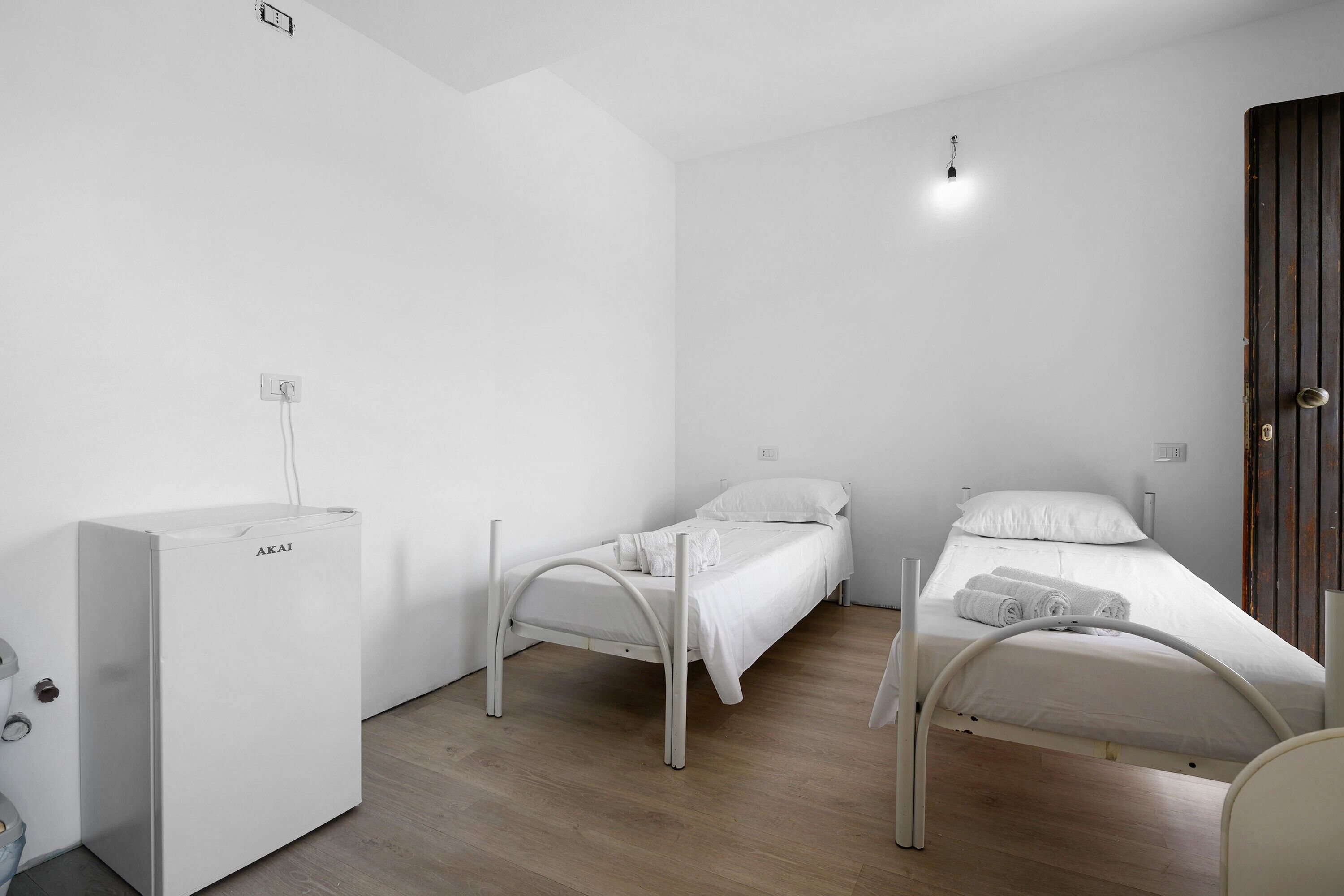 Een minimalistische kamer met twee eenpersoonsbedden, een klein wit kastje en een houten deur.