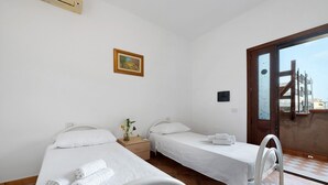 A hotel room with two single beds, a small table with a vase, a painting on the wall, and a view through a glass door. - Daluef Alloggi - Room 9 (Assemini)