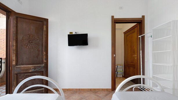 A room with a wooden door, a flat-screen TV mounted on the wall, and two white chairs with a table. - Daluef Alloggi - Room 9 (Assemini)