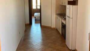 Un pasillo estrecho que conduce a un dormitorio con puerta, una zona de cocina con armarios y un refrigerador, y piso de baldosas.