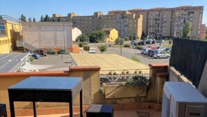 Una terraza en la azotea con mesa y sillas, un equipo de aire acondicionado y vistas a un estacionamiento y edificios a lo lejos.