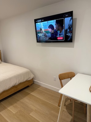 A room with a bed, a table, and a wall-mounted TV displaying a show.