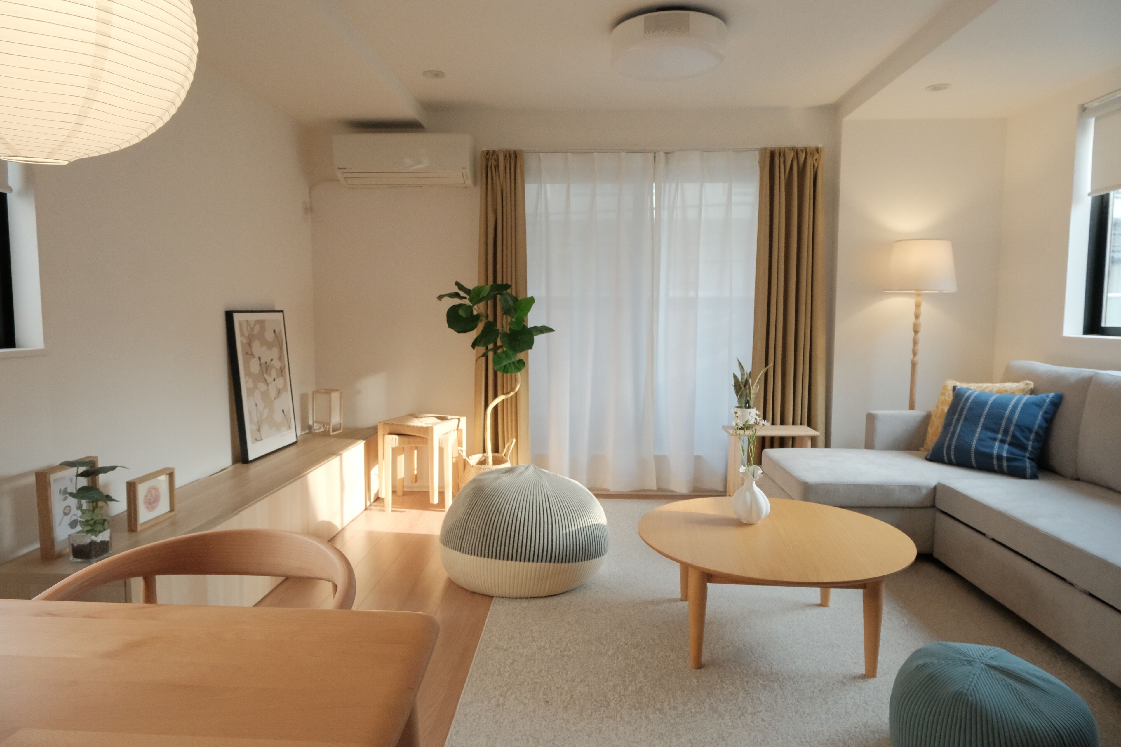 A modern living room with a sofa, a coffee table, a dining table, and a chair. There is a plant, a lamp, and a window with curtains.