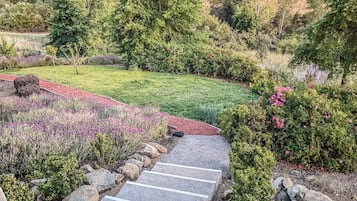 A landscaped garden with a stone pathway leading to a grassy area, surrounded by various shrubs and flowering plants.