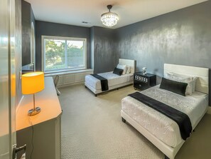 A hotel room with two beds, a desk with a lamp, a window with blinds, and a chandelier. - Serrano Village Villa (El Dorado Hills)