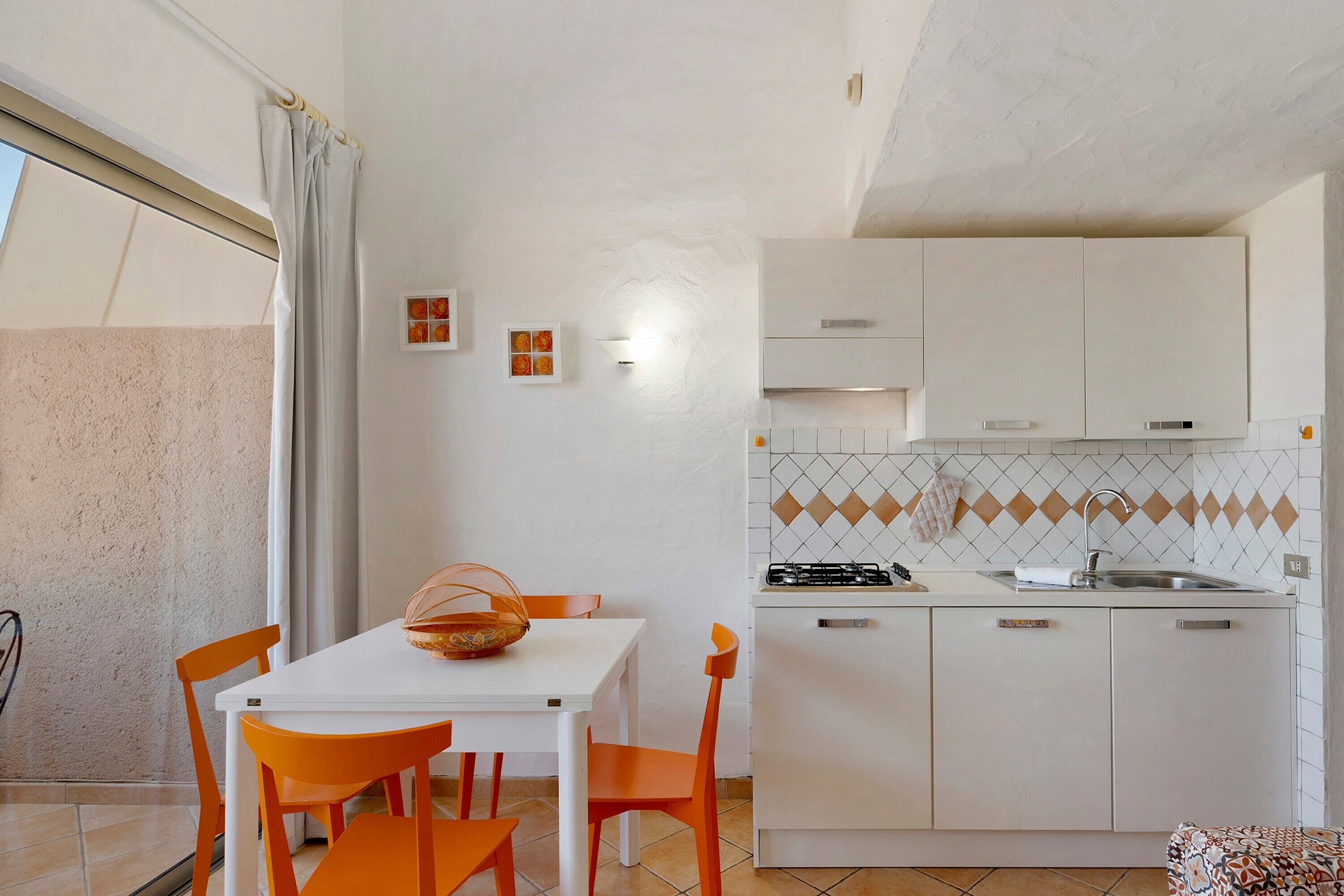 A compact kitchen with white cabinets, a gas stove, and a sink. There is a dining table with orange chairs and a basket on it.