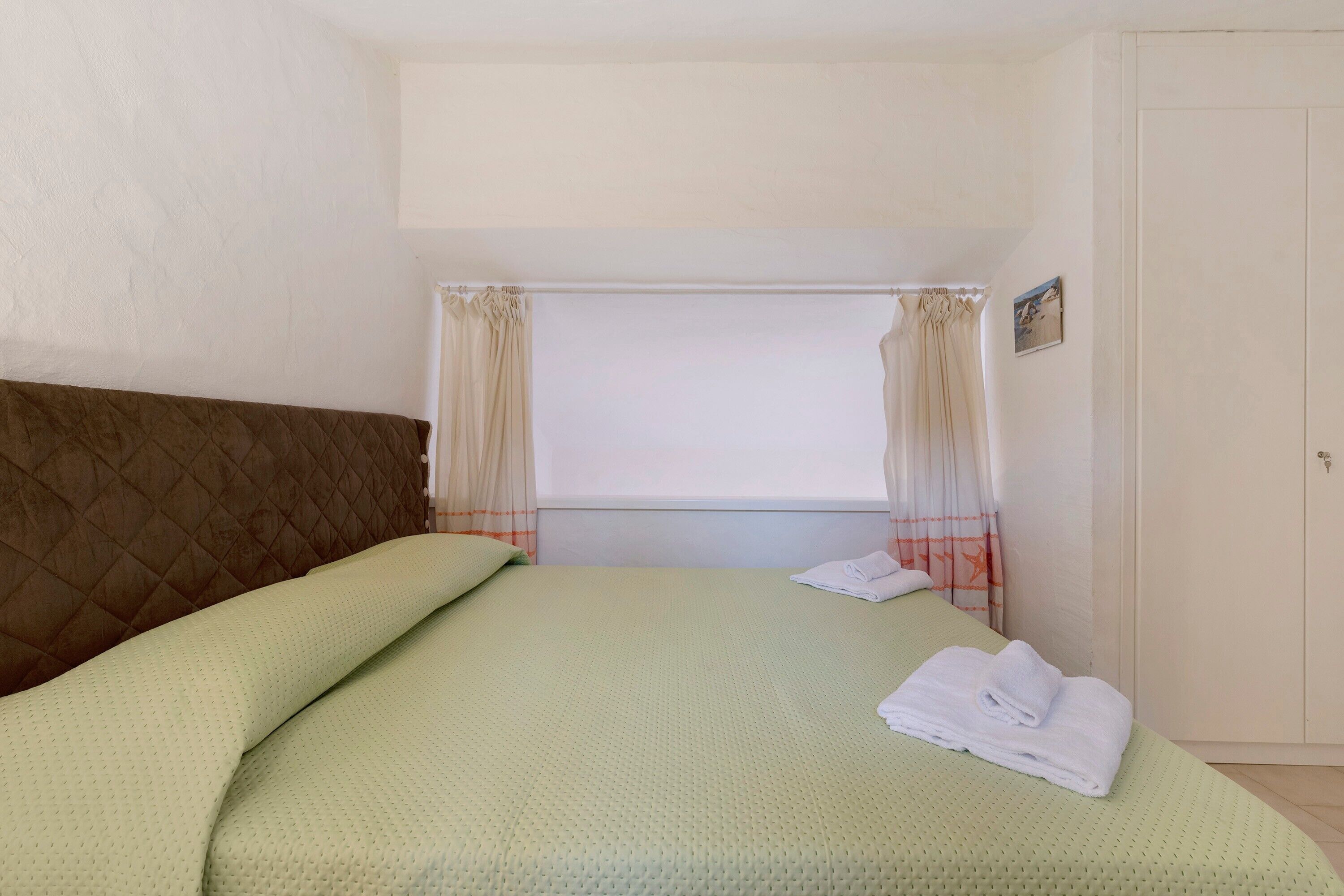 A single bed with a quilted headboard, a window with curtains, and a door with a keyhole.