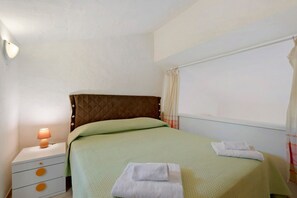 A single bed with a quilted headboard, a bedside table with a lamp, and a window with curtains. - Apartment 'Flat Pace E Mare' with Sea View, Shared Pool and Air Conditioning (Pulcheddu)