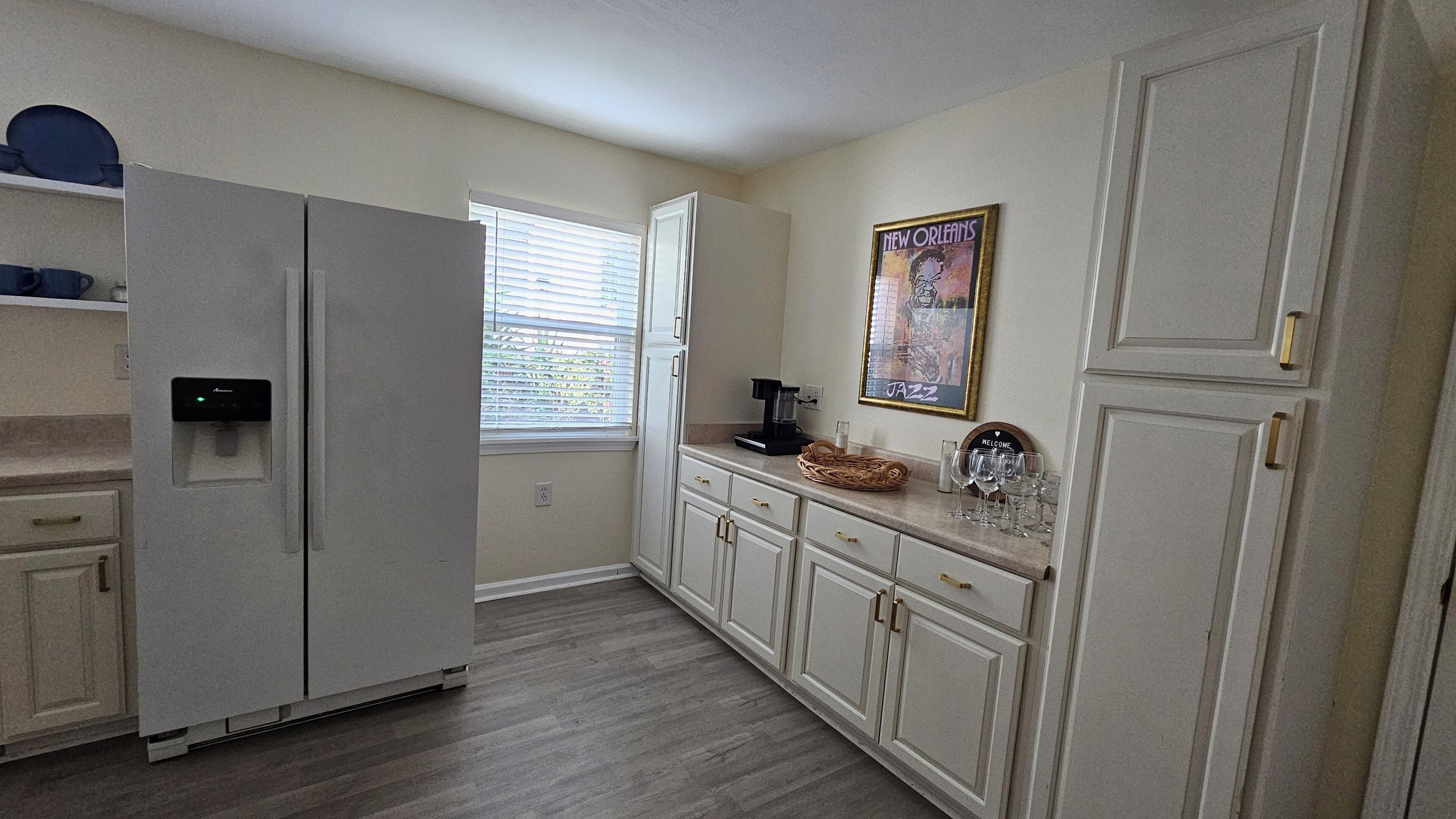 A kitchen with a refrigerator, cabinets, a coffee maker, and a window with blinds.