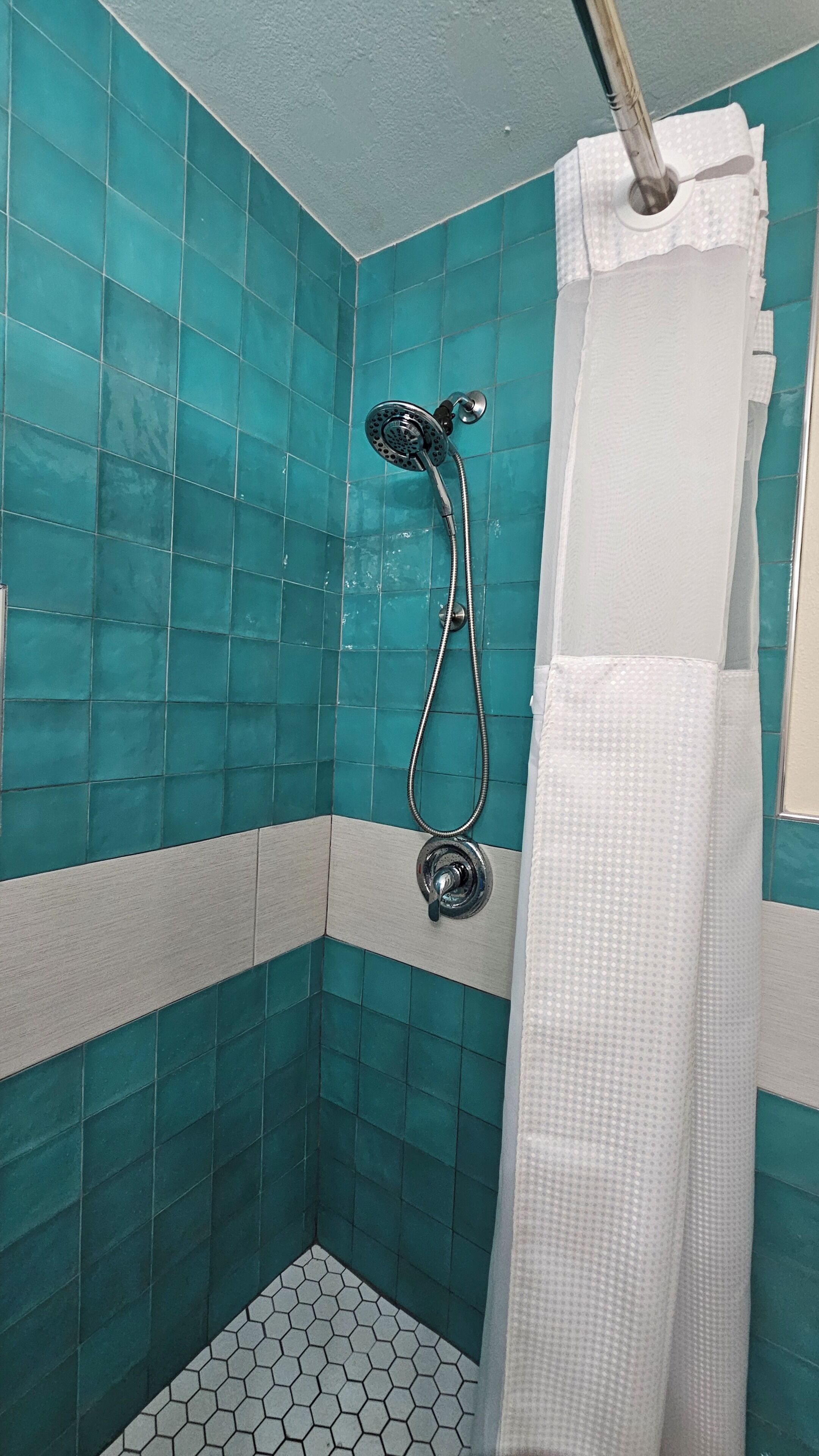 Une douche équipée d’un pommeau de douche à main, d’un rideau de douche blanc et d’un sol carrelé avec un motif hexagonal.
