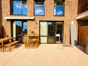 A two-story brick building with a patio area featuring a wooden table and benches, a sliding glass door leading to a kitchen, and a clear sky visible through the windows. - Bright and Spacious 2BDR Flat Westfield Stratford (Greater London)