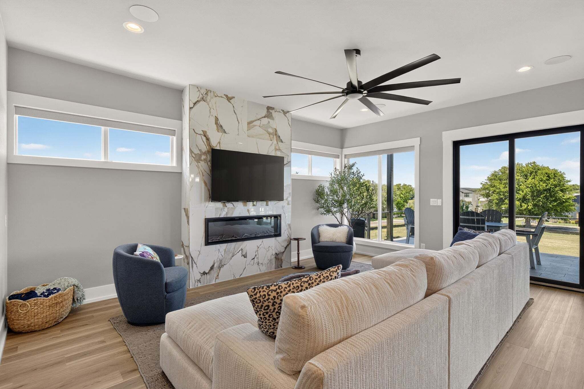 A spacious living room with a large sofa, a flat-screen TV mounted on a marble wall, a ceiling fan, and a view through a glass door to the outdoors.