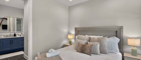 A bedroom with a bed, nightstands, a lamp, a mirror, and a bathroom with a sink and cabinets.