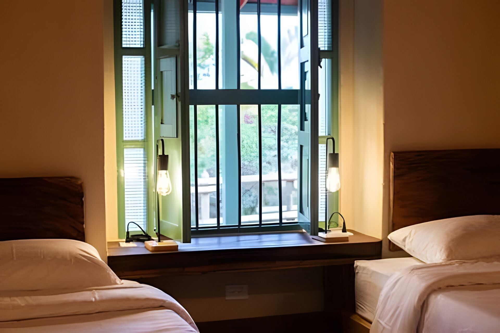 A bedroom with two beds, a window with blinds, and bedside lamps.