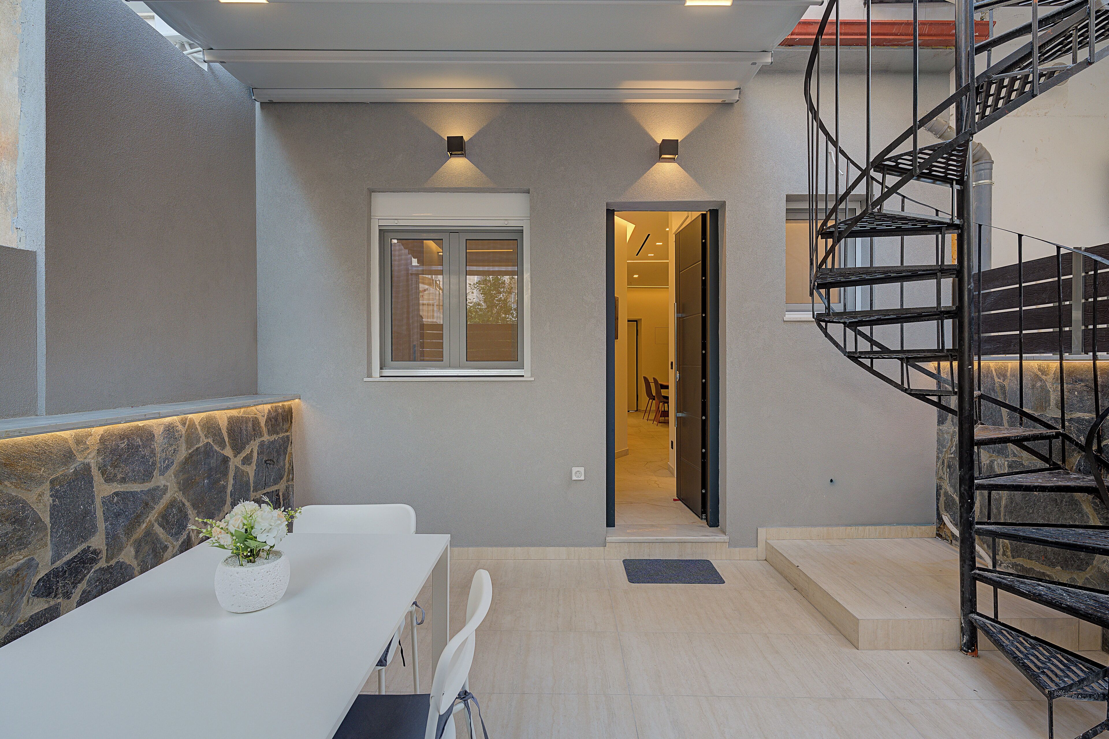 Un intérieur moderne avec un mur d’accent en pierre, une table blanche agrémentée d’une plante en pot, un escalier en colimaçon noir et une porte-fenêtre donnant sur une autre pièce.